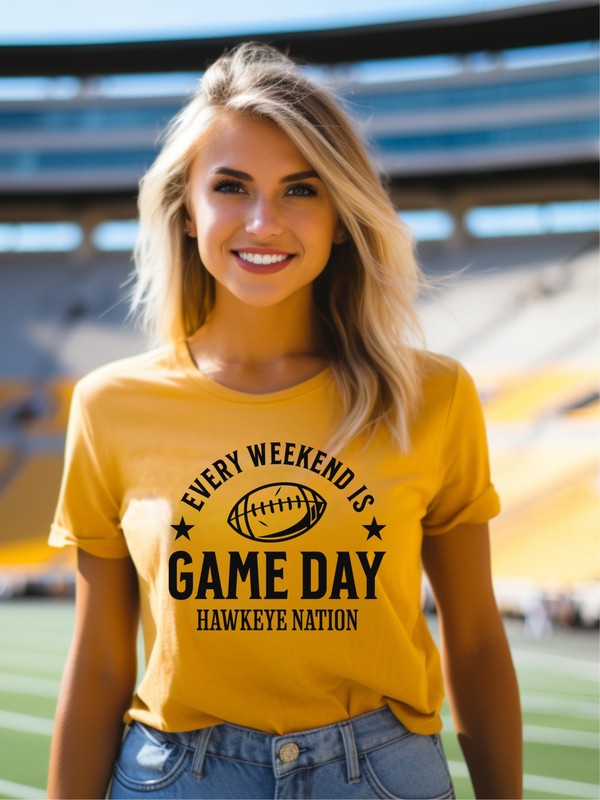 A Gameday Hawkeye Nation Graphic Crew Tee, featuring a yellow shirt with the text "Every Weekend is Game Day, Hawkeye Nation" and a football graphic, lies among jeans, a brown knit blanket, and autumn leaves.