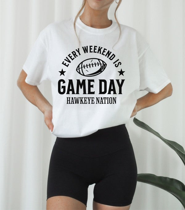 A Gameday Hawkeye Nation Graphic Crew Tee, featuring a yellow shirt with the text "Every Weekend is Game Day, Hawkeye Nation" and a football graphic, lies among jeans, a brown knit blanket, and autumn leaves.