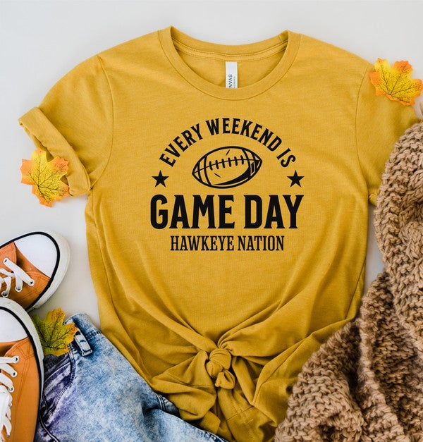 A Gameday Hawkeye Nation Graphic Crew Tee, featuring a yellow shirt with the text "Every Weekend is Game Day, Hawkeye Nation" and a football graphic, lies among jeans, a brown knit blanket, and autumn leaves.