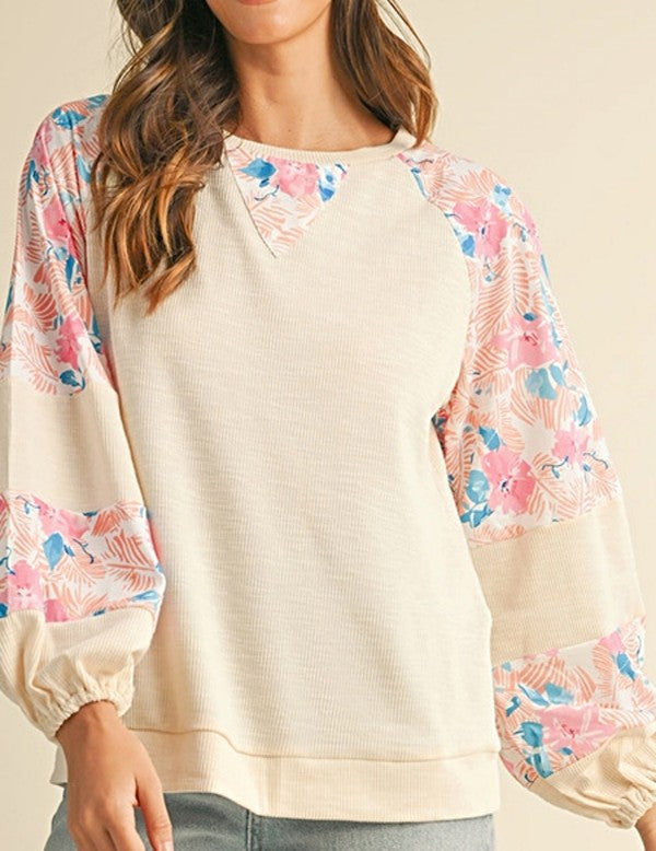 A person wearing the cotton Ribbed Floral Stitch Balloon Pullover, featuring cream tones and pink floral-patterned sleeves, stands near a snowy Christmas tree, exuding a subtle bohemian flair.