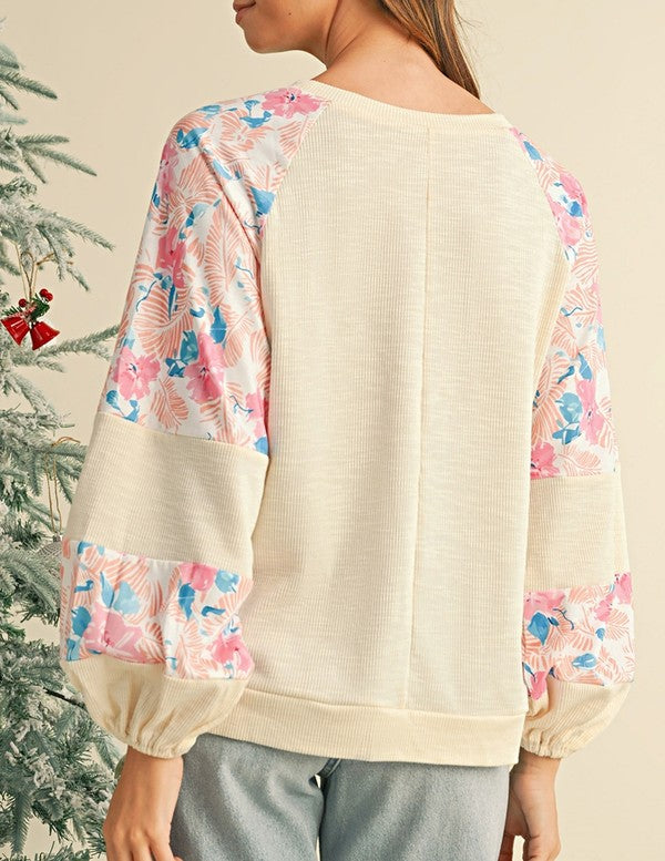 A person wearing the cotton Ribbed Floral Stitch Balloon Pullover, featuring cream tones and pink floral-patterned sleeves, stands near a snowy Christmas tree, exuding a subtle bohemian flair.