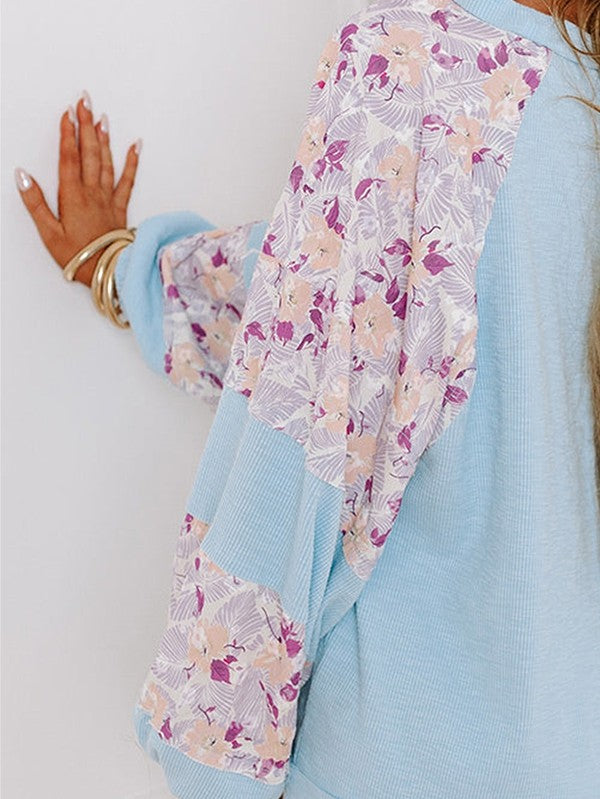 A person wearing the cotton Ribbed Floral Stitch Balloon Pullover, featuring cream tones and pink floral-patterned sleeves, stands near a snowy Christmas tree, exuding a subtle bohemian flair.