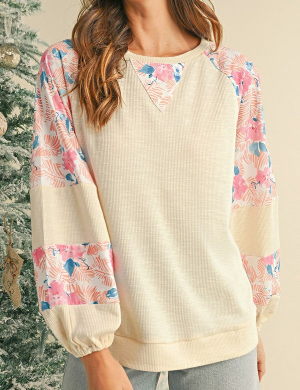 A person wearing the cotton Ribbed Floral Stitch Balloon Pullover, featuring cream tones and pink floral-patterned sleeves, stands near a snowy Christmas tree, exuding a subtle bohemian flair.