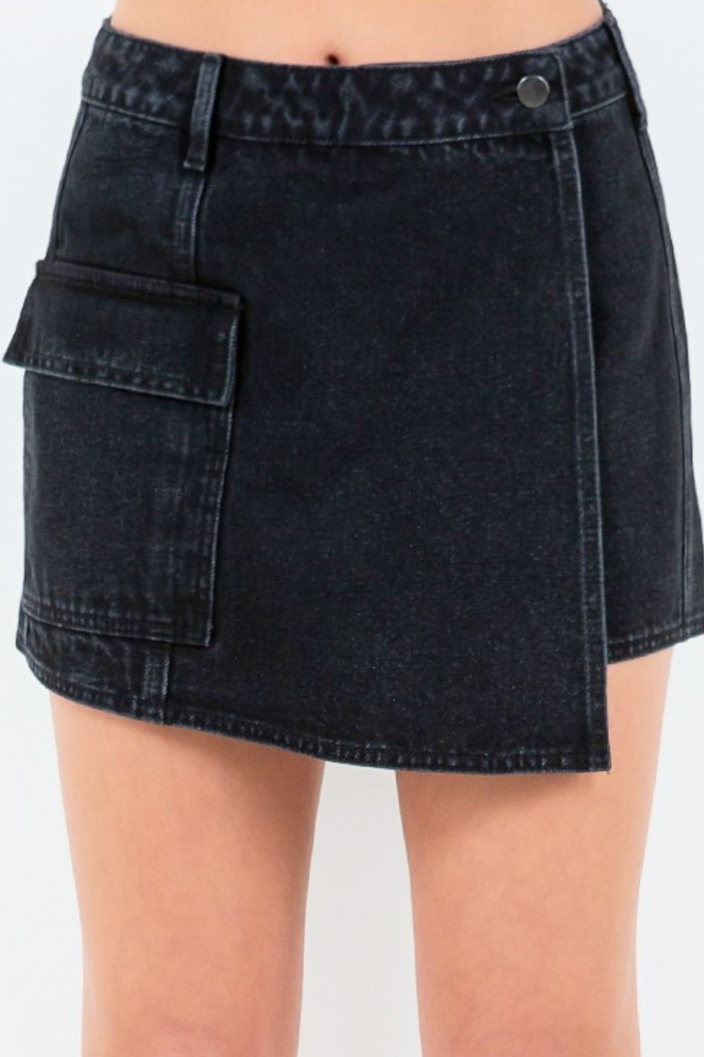 Front view of a person wearing an American Bazi Cargo Pocket Mini Skort in black denim with an asymmetrical front and button waist closure, paired with white sneakers.