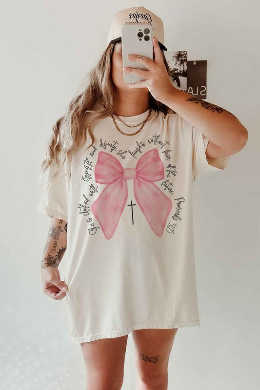 Person taking a mirror selfie wearing the PROVERBS PINK BOW CHRISTIAN OVERSIZED GRAPHIC TEE, made of premium cotton, featuring a large pink bow, a small cross below it, and cursive text around the design.