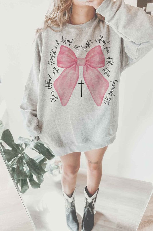 A person is wearing the PROVERBS PINK BOW CHRISTIAN OVERSIZED SWEATSHIRT made of premium cotton. The light gray design features a large pink bow with a black cross in the center and cursive text around it. Paired with cowboy boots, this Christian-inspired look blends comfort and style effortlessly.