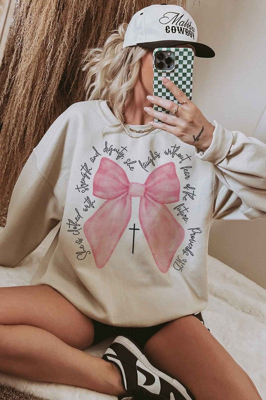 A person is wearing the PROVERBS PINK BOW CHRISTIAN OVERSIZED SWEATSHIRT made of premium cotton. The light gray design features a large pink bow with a black cross in the center and cursive text around it. Paired with cowboy boots, this Christian-inspired look blends comfort and style effortlessly.