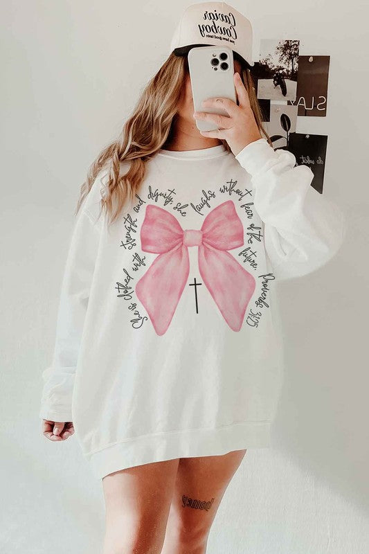 A person is wearing the PROVERBS PINK BOW CHRISTIAN OVERSIZED SWEATSHIRT made of premium cotton. The light gray design features a large pink bow with a black cross in the center and cursive text around it. Paired with cowboy boots, this Christian-inspired look blends comfort and style effortlessly.