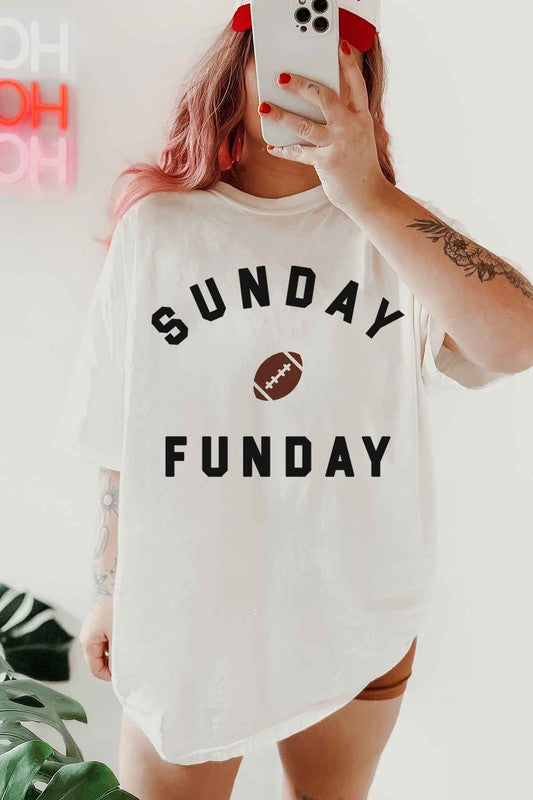 A person is wearing the SUNDAY FUN DAY FOOTBALL OVERSIZED GRAPHIC TEE made of premium cotton, featuring "Sunday Funday" and a small football graphic. They're holding a phone in front of their face, with part of a neon sign visible in the background—the perfect look for football game day.