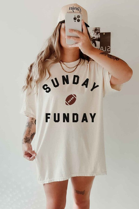 A person is wearing the SUNDAY FUN DAY FOOTBALL OVERSIZED GRAPHIC TEE made of premium cotton, featuring "Sunday Funday" and a small football graphic. They're holding a phone in front of their face, with part of a neon sign visible in the background—the perfect look for football game day.