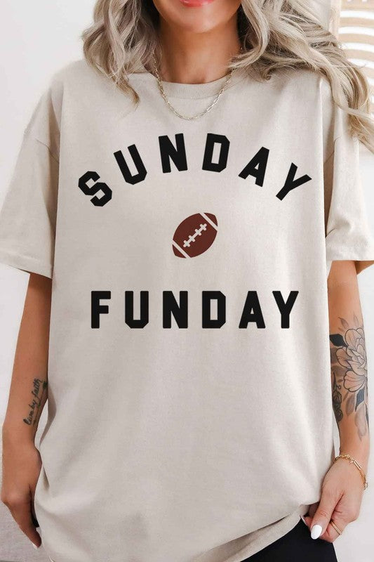 A person is wearing the SUNDAY FUN DAY FOOTBALL OVERSIZED GRAPHIC TEE made of premium cotton, featuring "Sunday Funday" and a small football graphic. They're holding a phone in front of their face, with part of a neon sign visible in the background—the perfect look for football game day.