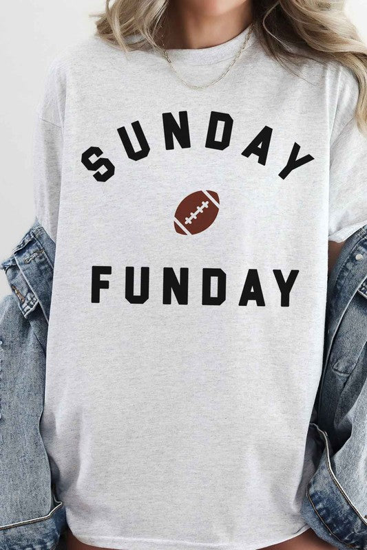 A person is wearing the SUNDAY FUN DAY FOOTBALL OVERSIZED GRAPHIC TEE made of premium cotton, featuring "Sunday Funday" and a small football graphic. They're holding a phone in front of their face, with part of a neon sign visible in the background—the perfect look for football game day.