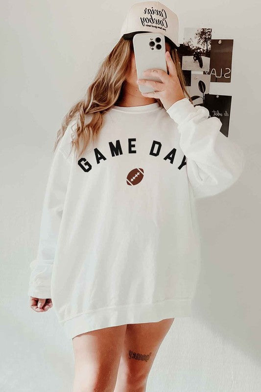 A person wearing a "GAME DAY FOOTBALL TAILGATE GRAPHIC SWEATSHIRT," black shorts, and a cap takes a mirror selfie with a phone, sitting on a white rug, ready for the tailgate festivities.