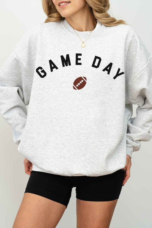 A person wearing a "GAME DAY FOOTBALL TAILGATE GRAPHIC SWEATSHIRT," black shorts, and a cap takes a mirror selfie with a phone, sitting on a white rug, ready for the tailgate festivities.