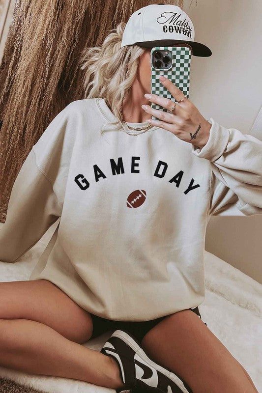 A person wearing a "GAME DAY FOOTBALL TAILGATE GRAPHIC SWEATSHIRT," black shorts, and a cap takes a mirror selfie with a phone, sitting on a white rug, ready for the tailgate festivities.