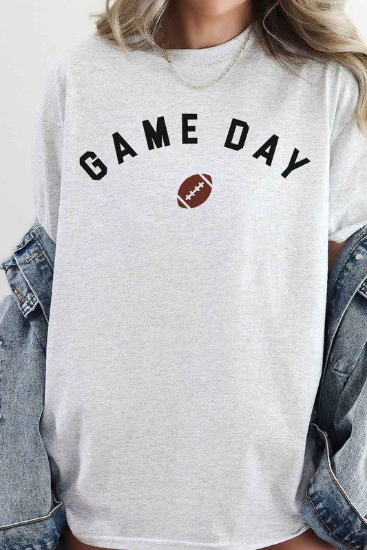 Person with pink hair wearing a GAME DAY FOOTBALL TAILGATE OVERSIZED GRAPHIC TEE, featuring "Game Day Football" and a football icon, taking a mirror selfie. Neon sign in the background.