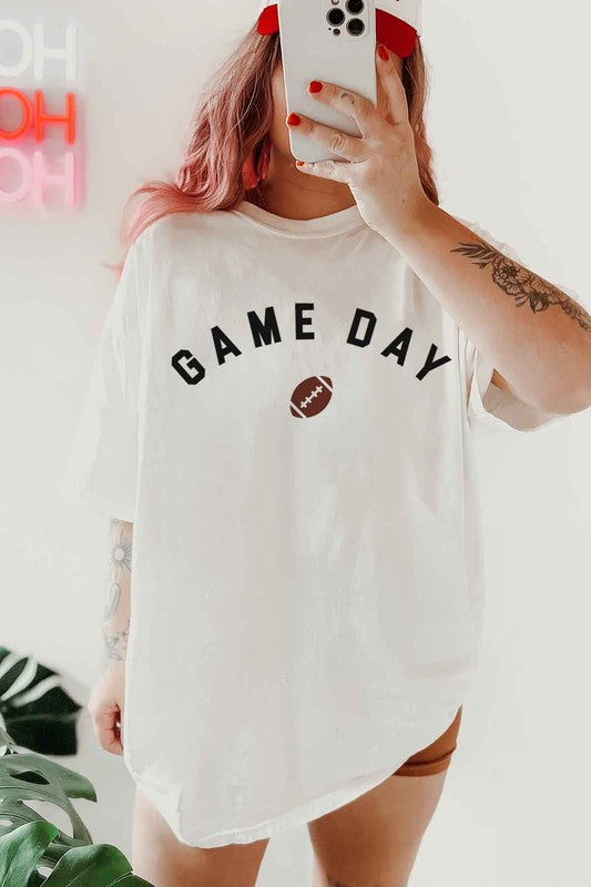 Person with pink hair wearing a GAME DAY FOOTBALL TAILGATE OVERSIZED GRAPHIC TEE, featuring "Game Day Football" and a football icon, taking a mirror selfie. Neon sign in the background.