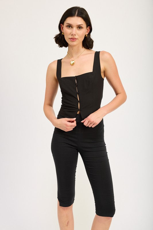 A woman with short dark hair stands against a plain background, wearing the "Sleeveless Top with Button Up Detail," made from imported polyester and featuring front buttons. She pairs it with knee-length pants and a heart-shaped pendant necklace, maintaining a neutral expression.