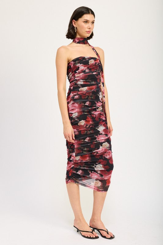 A woman wearing the SHIRRED FLORAL MINI DRESS WITH SCARF, a sleeveless, floral-print, form-fitting dress made of polyester, with a matching neck sash. She stands with one hand on her hip and pairs the outfit with black sandals. Available in size small.