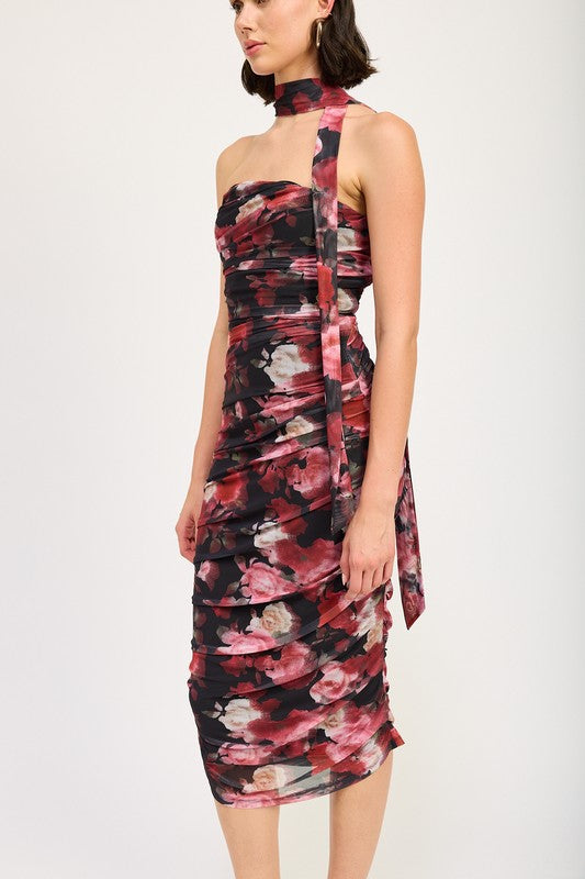 A woman wearing the SHIRRED FLORAL MINI DRESS WITH SCARF, a sleeveless, floral-print, form-fitting dress made of polyester, with a matching neck sash. She stands with one hand on her hip and pairs the outfit with black sandals. Available in size small.