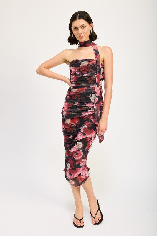 A woman wearing the SHIRRED FLORAL MINI DRESS WITH SCARF, a sleeveless, floral-print, form-fitting dress made of polyester, with a matching neck sash. She stands with one hand on her hip and pairs the outfit with black sandals. Available in size small.