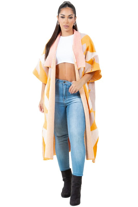 A woman is wearing the SEXY FASHION SWEATER CARDIGAN, a longline, orange and white patterned open-front cardigan with knit 3/4 sleeves, paired with a white crop top, blue jeans, and black boots.