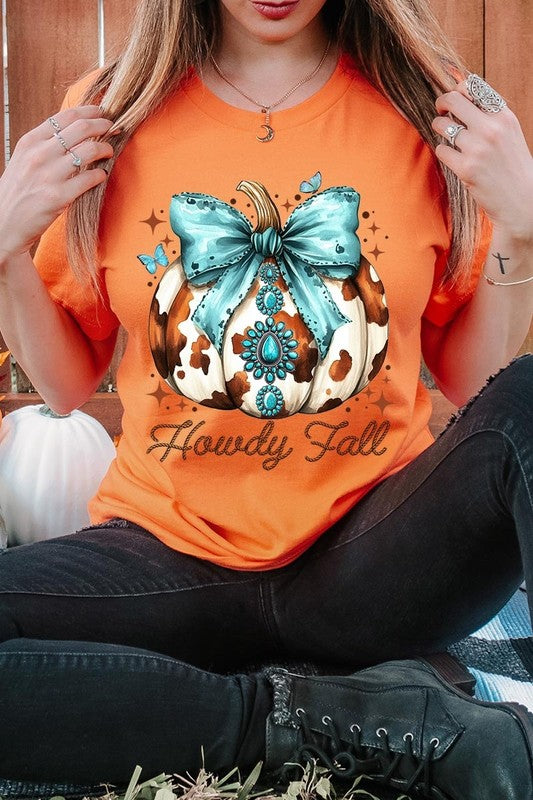 Person wearing the Howdy Fall Western Pumpkin Graphic Tee, featuring an illustrated pumpkin adorned with a blue bow and turquoise jewelry, along with the text "Howdy Fall." This beige unisex crew neck shirt pairs perfectly with their denim jacket.