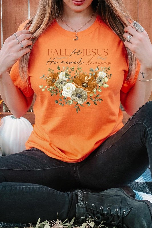 A person wearing the "Fall For Jesus He Never Leaves Graphic Tee," which is a dark blue, unisex crew neck featuring a floral design and the text "FALL FOR JESUS He never leaves," sits cross-legged, holding the sides of the shirt. The graphic tee is perfectly paired with light-wash jeans.