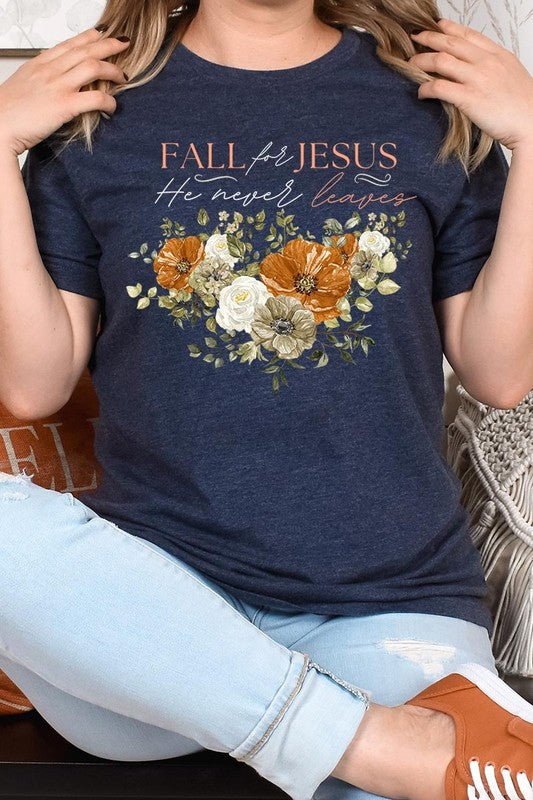 A person wearing the "Fall For Jesus He Never Leaves Graphic Tee," which is a dark blue, unisex crew neck featuring a floral design and the text "FALL FOR JESUS He never leaves," sits cross-legged, holding the sides of the shirt. The graphic tee is perfectly paired with light-wash jeans.