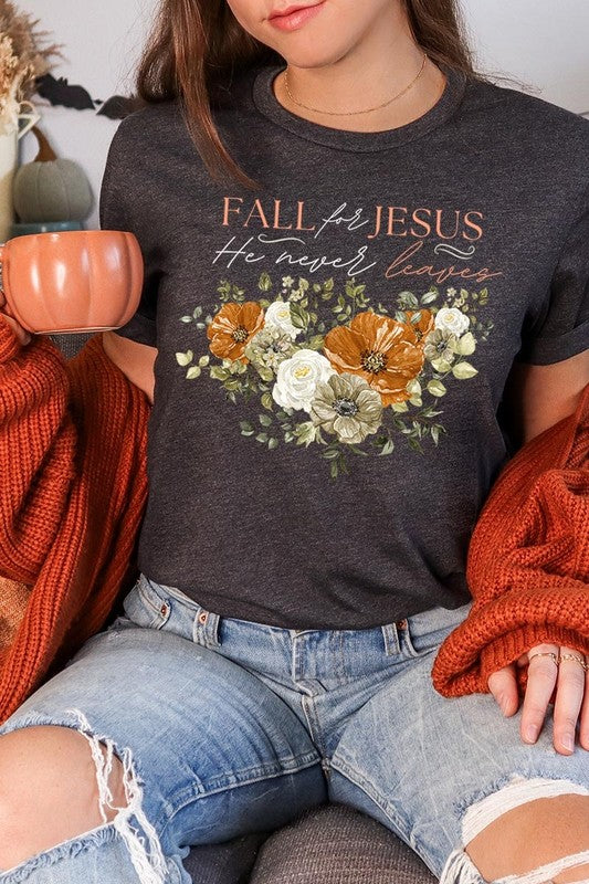 A person wearing the "Fall For Jesus He Never Leaves Graphic Tee," which is a dark blue, unisex crew neck featuring a floral design and the text "FALL FOR JESUS He never leaves," sits cross-legged, holding the sides of the shirt. The graphic tee is perfectly paired with light-wash jeans.