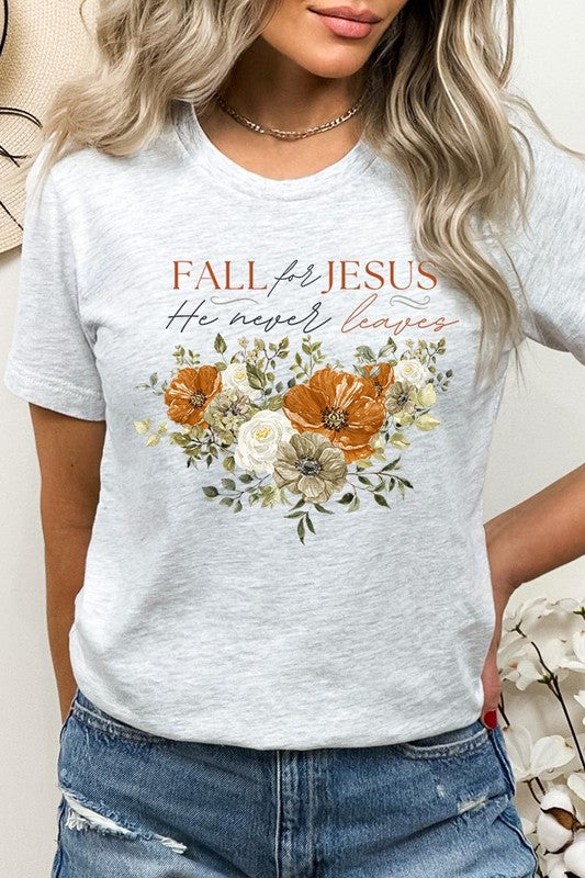 A person wearing the "Fall For Jesus He Never Leaves Graphic Tee," which is a dark blue, unisex crew neck featuring a floral design and the text "FALL FOR JESUS He never leaves," sits cross-legged, holding the sides of the shirt. The graphic tee is perfectly paired with light-wash jeans.