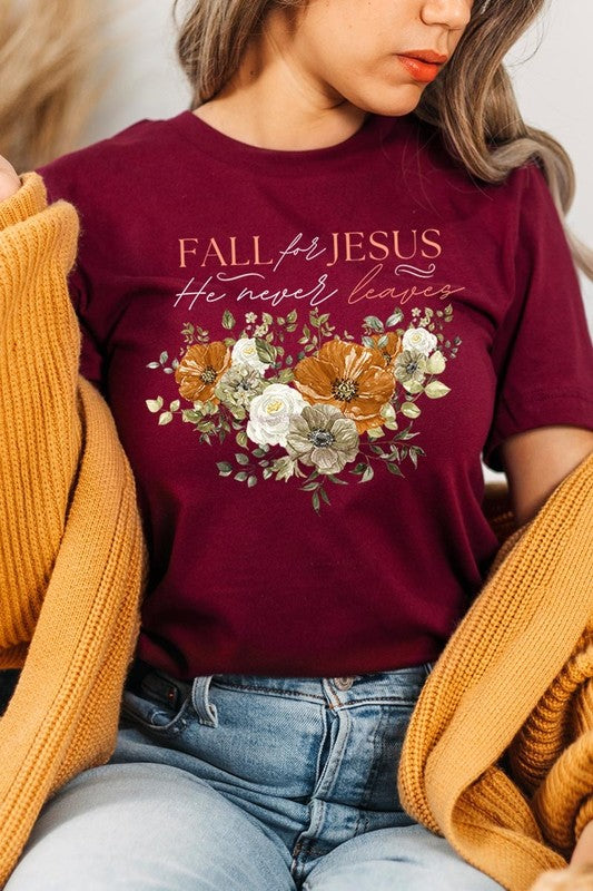 A person wearing the "Fall For Jesus He Never Leaves Graphic Tee," which is a dark blue, unisex crew neck featuring a floral design and the text "FALL FOR JESUS He never leaves," sits cross-legged, holding the sides of the shirt. The graphic tee is perfectly paired with light-wash jeans.