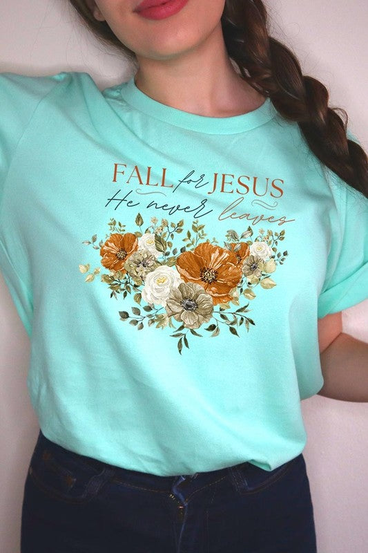 A person wearing the "Fall For Jesus He Never Leaves Graphic Tee," which is a dark blue, unisex crew neck featuring a floral design and the text "FALL FOR JESUS He never leaves," sits cross-legged, holding the sides of the shirt. The graphic tee is perfectly paired with light-wash jeans.