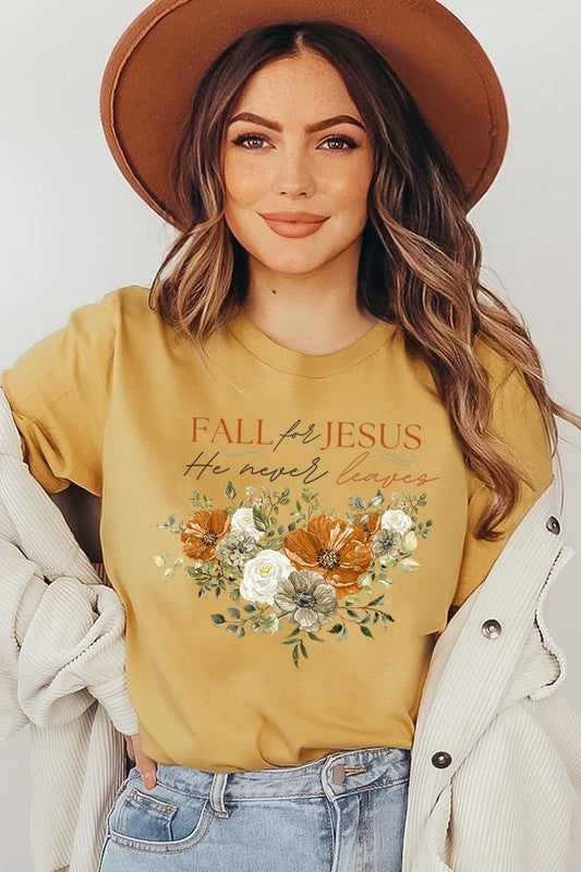 A person wearing the "Fall For Jesus He Never Leaves Graphic Tee," which is a dark blue, unisex crew neck featuring a floral design and the text "FALL FOR JESUS He never leaves," sits cross-legged, holding the sides of the shirt. The graphic tee is perfectly paired with light-wash jeans.