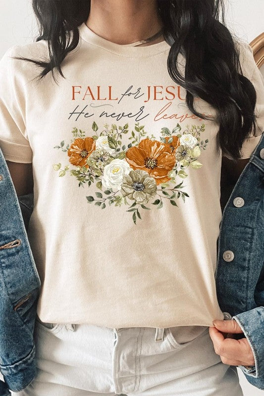 A person wearing the "Fall For Jesus He Never Leaves Graphic Tee," which is a dark blue, unisex crew neck featuring a floral design and the text "FALL FOR JESUS He never leaves," sits cross-legged, holding the sides of the shirt. The graphic tee is perfectly paired with light-wash jeans.