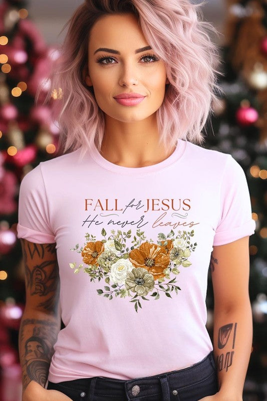 A person wearing the "Fall For Jesus He Never Leaves Graphic Tee," which is a dark blue, unisex crew neck featuring a floral design and the text "FALL FOR JESUS He never leaves," sits cross-legged, holding the sides of the shirt. The graphic tee is perfectly paired with light-wash jeans.