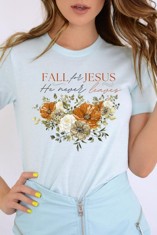 A person wearing the "Fall For Jesus He Never Leaves Graphic Tee," which is a dark blue, unisex crew neck featuring a floral design and the text "FALL FOR JESUS He never leaves," sits cross-legged, holding the sides of the shirt. The graphic tee is perfectly paired with light-wash jeans.