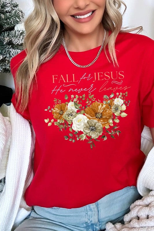 A person wearing the "Fall For Jesus He Never Leaves Graphic Tee," which is a dark blue, unisex crew neck featuring a floral design and the text "FALL FOR JESUS He never leaves," sits cross-legged, holding the sides of the shirt. The graphic tee is perfectly paired with light-wash jeans.