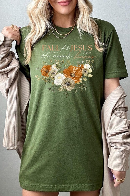 A person wearing the "Fall For Jesus He Never Leaves Graphic Tee," which is a dark blue, unisex crew neck featuring a floral design and the text "FALL FOR JESUS He never leaves," sits cross-legged, holding the sides of the shirt. The graphic tee is perfectly paired with light-wash jeans.