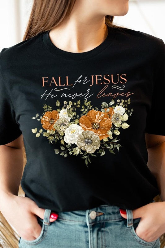 A person wearing the "Fall For Jesus He Never Leaves Graphic Tee," which is a dark blue, unisex crew neck featuring a floral design and the text "FALL FOR JESUS He never leaves," sits cross-legged, holding the sides of the shirt. The graphic tee is perfectly paired with light-wash jeans.