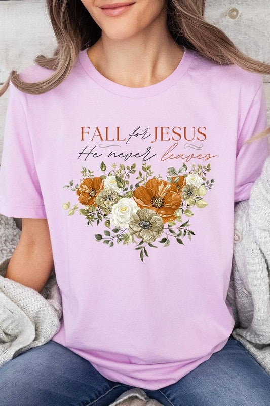 A person wearing the "Fall For Jesus He Never Leaves Graphic Tee," which is a dark blue, unisex crew neck featuring a floral design and the text "FALL FOR JESUS He never leaves," sits cross-legged, holding the sides of the shirt. The graphic tee is perfectly paired with light-wash jeans.