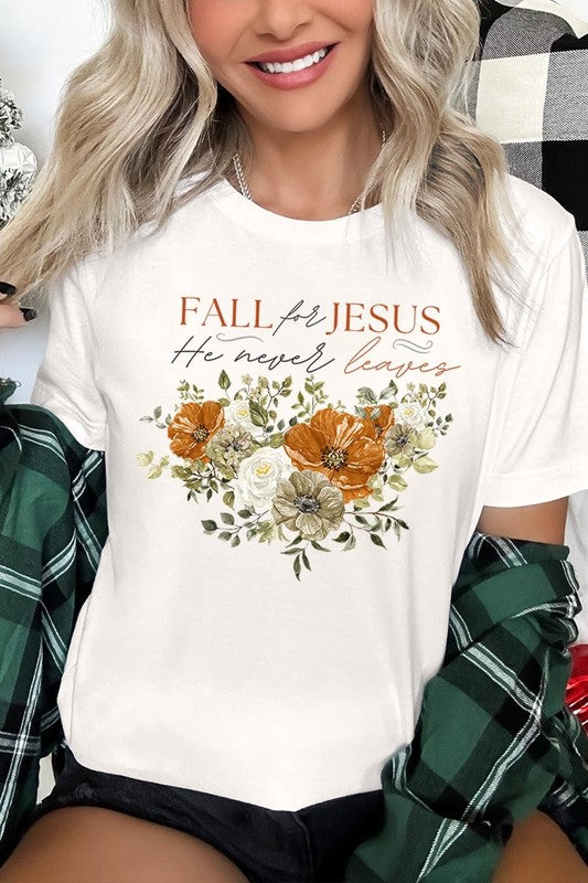 A person wearing the "Fall For Jesus He Never Leaves Graphic Tee," which is a dark blue, unisex crew neck featuring a floral design and the text "FALL FOR JESUS He never leaves," sits cross-legged, holding the sides of the shirt. The graphic tee is perfectly paired with light-wash jeans.