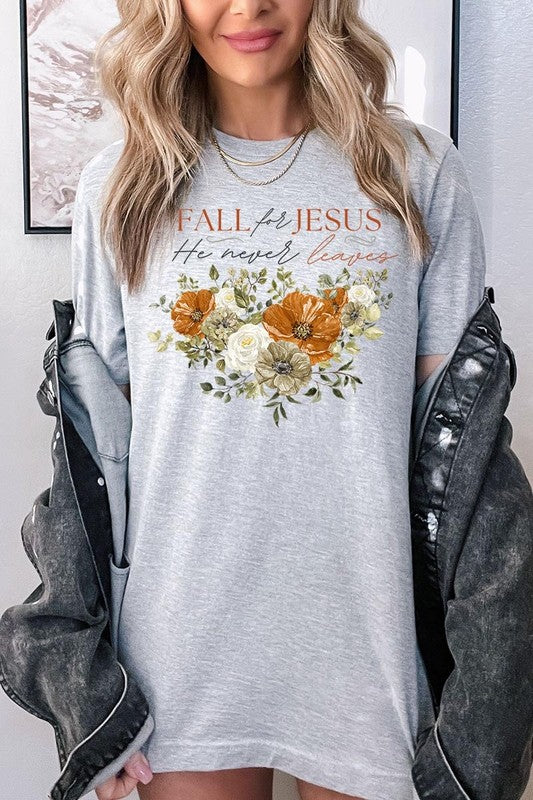 A person wearing the "Fall For Jesus He Never Leaves Graphic Tee," which is a dark blue, unisex crew neck featuring a floral design and the text "FALL FOR JESUS He never leaves," sits cross-legged, holding the sides of the shirt. The graphic tee is perfectly paired with light-wash jeans.