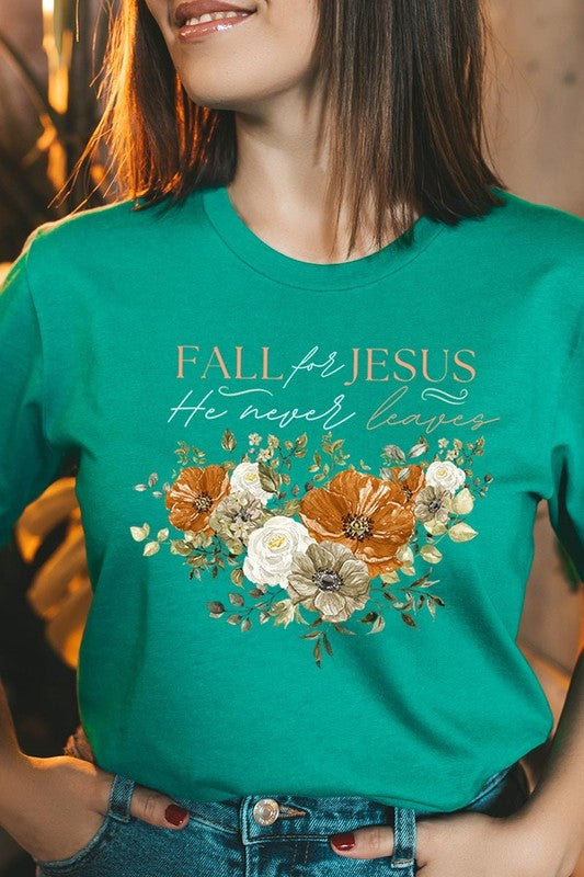 A person wearing the "Fall For Jesus He Never Leaves Graphic Tee," which is a dark blue, unisex crew neck featuring a floral design and the text "FALL FOR JESUS He never leaves," sits cross-legged, holding the sides of the shirt. The graphic tee is perfectly paired with light-wash jeans.