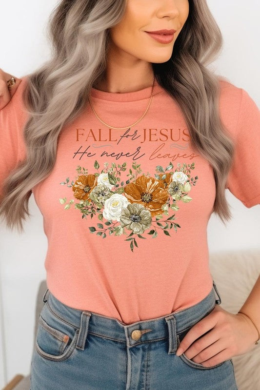 A person wearing the "Fall For Jesus He Never Leaves Graphic Tee," which is a dark blue, unisex crew neck featuring a floral design and the text "FALL FOR JESUS He never leaves," sits cross-legged, holding the sides of the shirt. The graphic tee is perfectly paired with light-wash jeans.