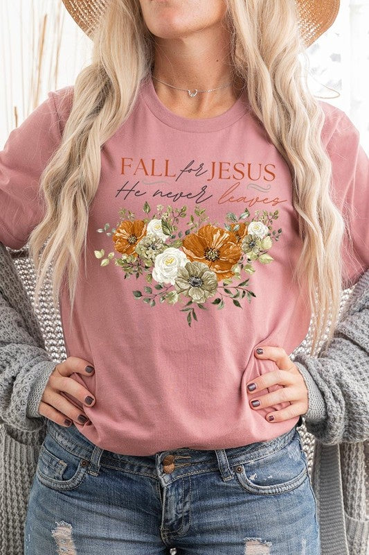 A person wearing the "Fall For Jesus He Never Leaves Graphic Tee," which is a dark blue, unisex crew neck featuring a floral design and the text "FALL FOR JESUS He never leaves," sits cross-legged, holding the sides of the shirt. The graphic tee is perfectly paired with light-wash jeans.