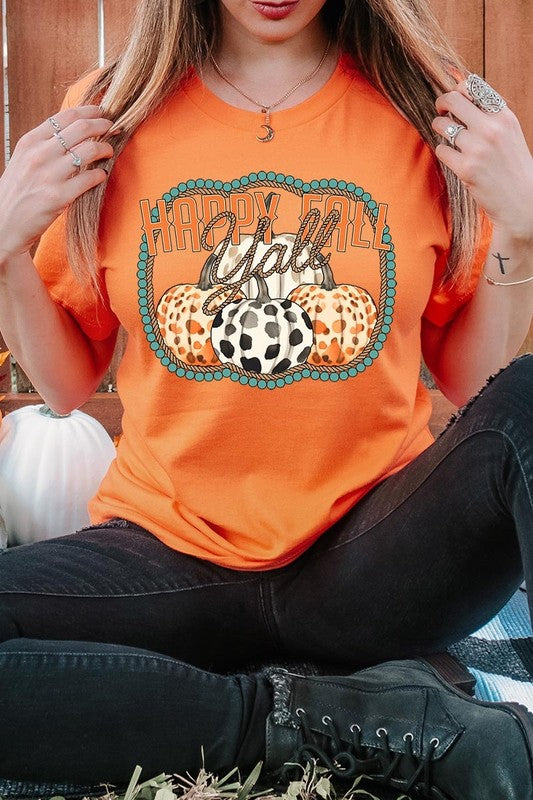 A person wearing a top-quality "Happy Fall Y'all" Graphic Tee featuring a design with three patterned pumpkins, paired with jeans and an orange cardigan, holds a pumpkin-shaped mug.