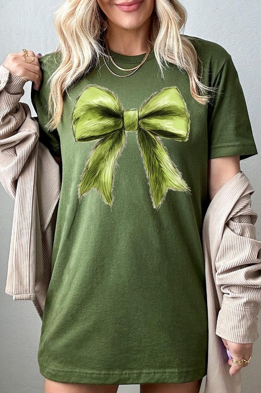 A person wearing a white Grinchmas Coquette Bow Graphic Tee with a green plaid shirt tied loosely over their shoulders. Only the lower part of their face is visible.