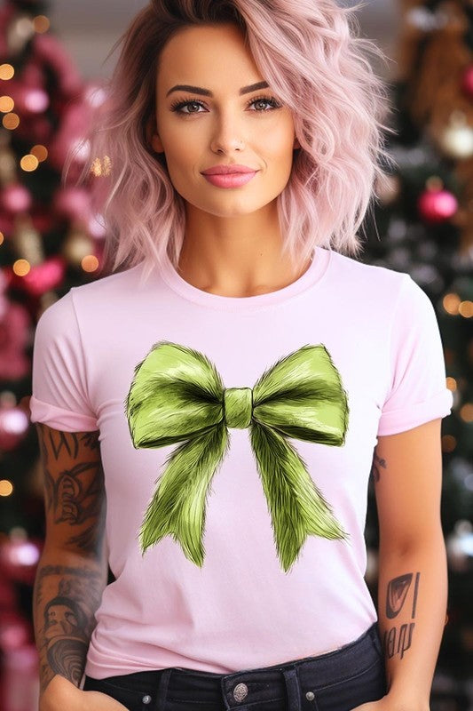 A person wearing a white Grinchmas Coquette Bow Graphic Tee with a green plaid shirt tied loosely over their shoulders. Only the lower part of their face is visible.