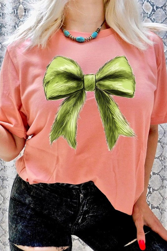 A person wearing a white Grinchmas Coquette Bow Graphic Tee with a green plaid shirt tied loosely over their shoulders. Only the lower part of their face is visible.